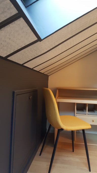 Une chaise jaune placée contre un mur sous un toit mansardé avec un puits de lumière.