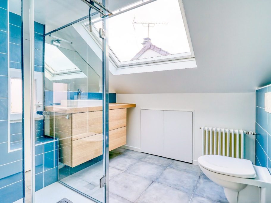 Salle de bains sous combles après travaux à Chelles salle de bain sous combles-murs blancs, douche en carrelage bleu, meuble vasque à deux tiroirs en bois clair. WC suspendu en face.