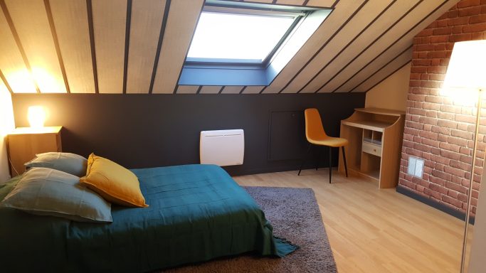 Chambre mansardée avec un lit, un bureau, et une fenêtre de toit. Deco moderne et chaleureuse.