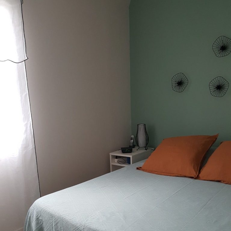 Chambre avec un lit, coussins orange, mur vert et fenêtres drapées de blanc.