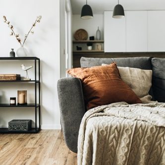 Salon lumineux avec canapé gris, coussins orange, couverture beige et étagère décorative.