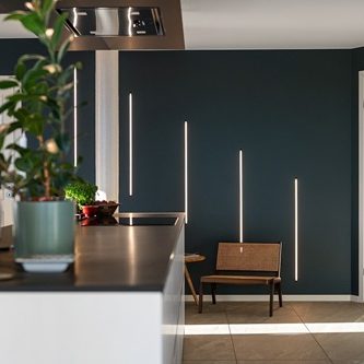 proposition d’aménagement intérieur d’une cuisine Mur sombre avec des bandes lumineuses verticales et un banc en osier. Plante en pot sur le comptoir.